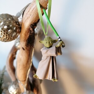 Puede incluir: Adorno navide&ntilde;o de Yoda con un sable de luz verde, colgado de una cinta verde. La figura de Yoda es verde y beige, con una t&uacute;nica beige. El adorno cuelga de madera flotante, con bolas plateadas en el fondo.