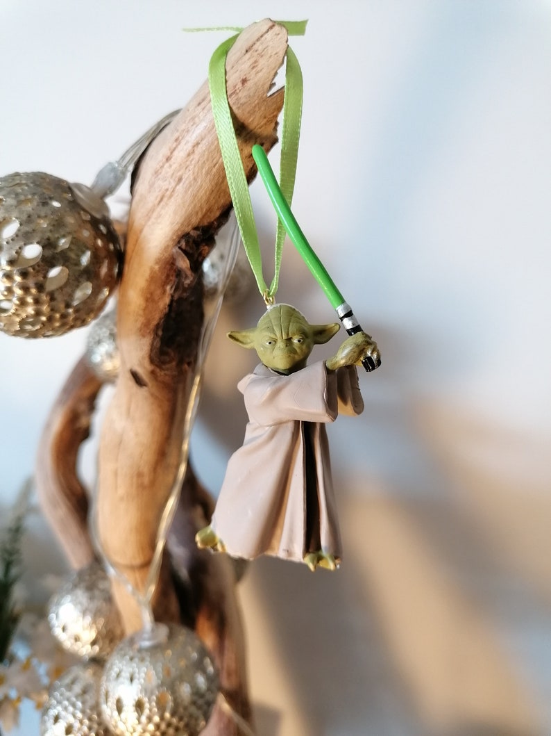 Puede incluir: Adorno navide&ntilde;o de Yoda con un sable de luz verde, colgado de una cinta verde. La figura de Yoda es verde y beige, con una t&uacute;nica beige. El adorno cuelga de madera flotante, con bolas plateadas en el fondo.