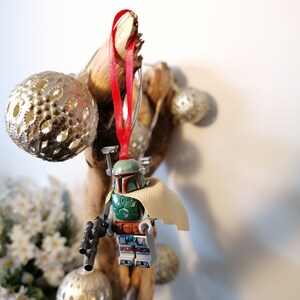 May include: A green and brown Lego Star Wars Boba Fett minifigure ornament with a red ribbon hanging from a branch with silver ornaments.