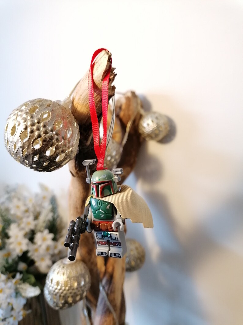 May include: A green and brown Lego Star Wars Boba Fett minifigure ornament with a red ribbon hanging from a branch with silver ornaments.