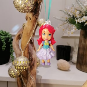 Peut inclure: Une petite poup&eacute;e en plastique repr&eacute;sentant une princesse aux cheveux roux, v&ecirc;tue d'une robe violette et d'une couronne argent&eacute;e, suspendue &agrave; un ruban turquoise sur une branche de bois flott&eacute; avec des ornements dor&eacute;s.
