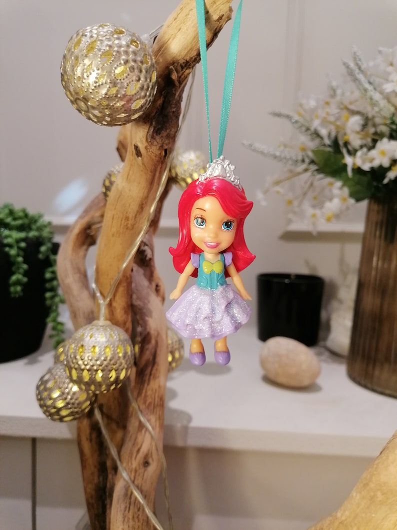 Peut inclure: Une petite poup&eacute;e en plastique repr&eacute;sentant une princesse aux cheveux roux, v&ecirc;tue d'une robe violette et d'une couronne argent&eacute;e, suspendue &agrave; un ruban turquoise sur une branche de bois flott&eacute; avec des ornements dor&eacute;s.