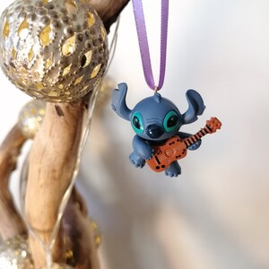 Op de afbeelding: Blauw Stitch-personage ornament dat een bruine gitaar bespeelt, hangend aan een paars lint.