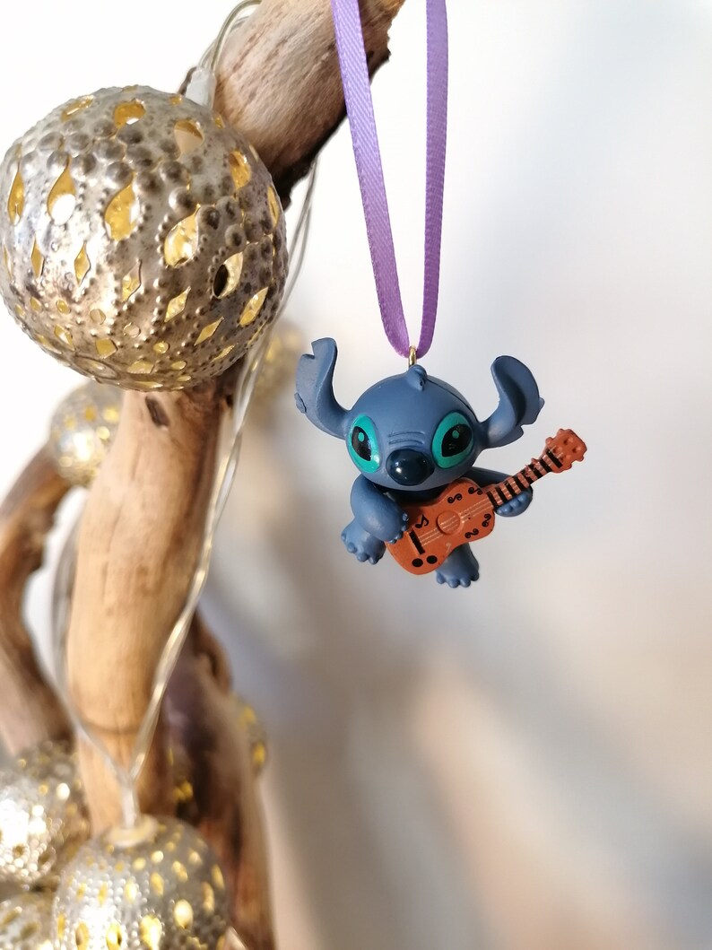 Op de afbeelding: Blauw Stitch-personage ornament dat een bruine gitaar bespeelt, hangend aan een paars lint.