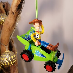 May include: A green toy car with blue flames and a figure of Woody from Toy Story driving it. The car is hanging from a green ribbon.