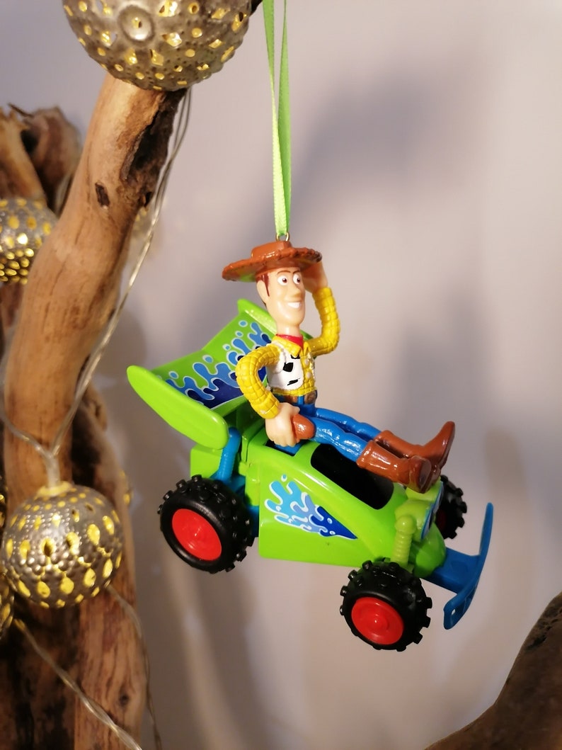 May include: A green toy car with blue flames and a figure of Woody from Toy Story driving it. The car is hanging from a green ribbon.