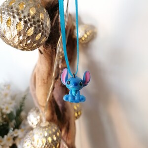 May include: A blue Stitch character charm hangs from a turquoise ribbon. The charm is a 3D representation of the popular Disney character.