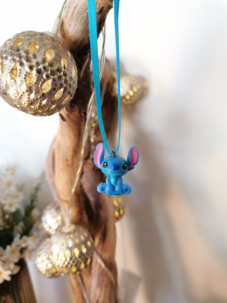 May include: A blue Stitch character charm hangs from a turquoise ribbon. The charm is a 3D representation of the popular Disney character.