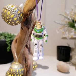 May include: A green and white Buzz Lightyear action figure ornament hanging from a purple ribbon. The ornament is attached to a piece of driftwood with string lights.