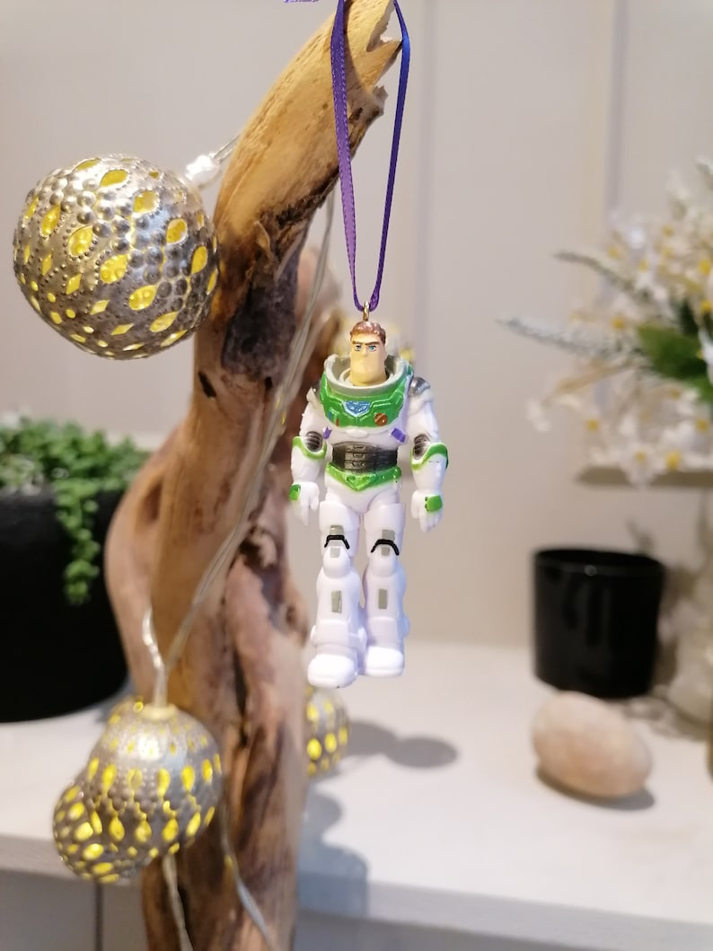 May include: A green and white Buzz Lightyear action figure ornament hanging from a purple ribbon. The ornament is attached to a piece of driftwood with string lights.