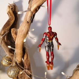 May include: A red and silver Iron Man Christmas ornament hanging from a branch with a red ribbon. The ornament is detailed with a metallic finish and features a classic Iron Man design.