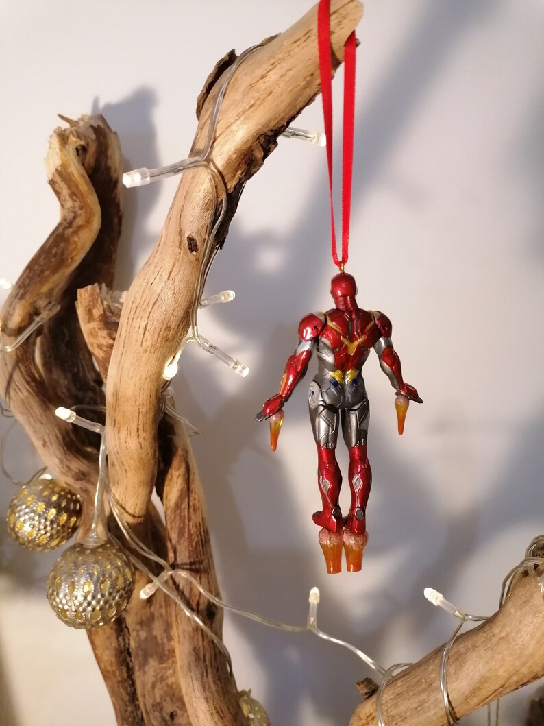 May include: A red and silver Iron Man Christmas ornament hanging from a branch with a red ribbon. The ornament is detailed with a metallic finish and features a classic Iron Man design.