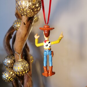 May include: A red string ornament featuring a plastic toy figurine of Woody from the Toy Story franchise. Woody is wearing his signature brown hat, blue jeans, and a yellow vest with a red bandana. He is holding his right arm up in the air, with his left arm bent at the elbow.