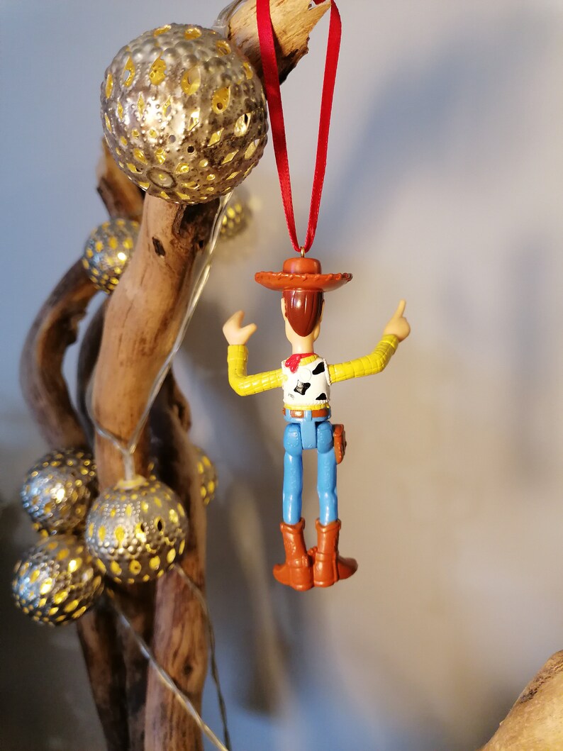 May include: A red string ornament featuring a plastic toy figurine of Woody from the Toy Story franchise. Woody is wearing his signature brown hat, blue jeans, and a yellow vest with a red bandana. He is holding his right arm up in the air, with his left arm bent at the elbow.