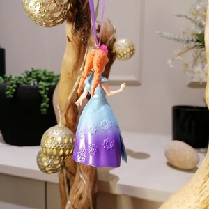 K&ouml;nnte beinhalten: Ein lila und blauer Plastik-Anh&auml;nger einer M&auml;dchenfigur mit langen roten Haaren, die ein Kleid mit Schneeflockenmuster tr&auml;gt. Der Anh&auml;nger h&auml;ngt an einem lila Band.