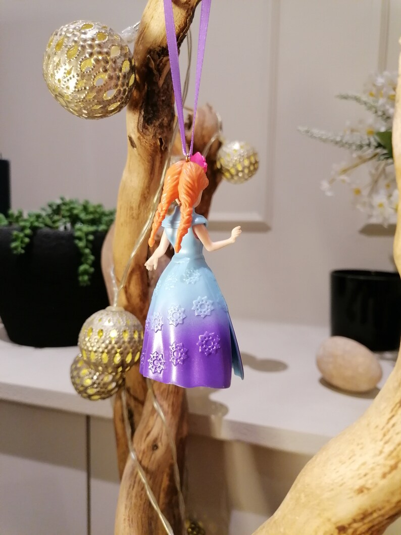 K&ouml;nnte beinhalten: Ein lila und blauer Plastik-Anh&auml;nger einer M&auml;dchenfigur mit langen roten Haaren, die ein Kleid mit Schneeflockenmuster tr&auml;gt. Der Anh&auml;nger h&auml;ngt an einem lila Band.