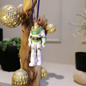 May include: A white and green plastic figurine of Buzz Lightyear from the Toy Story movies hanging from a purple ribbon on a piece of driftwood.