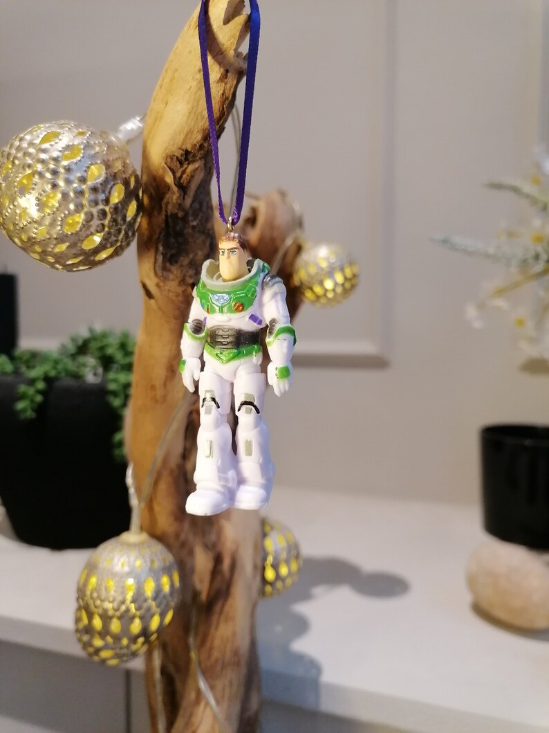 May include: A white and green plastic figurine of Buzz Lightyear from the Toy Story movies hanging from a purple ribbon on a piece of driftwood.