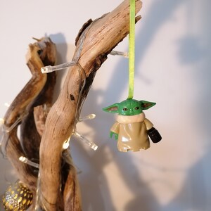May include: A green and brown Yoda ornament hanging from a branch with a green ribbon. The ornament is made of plastic and has a detailed design.