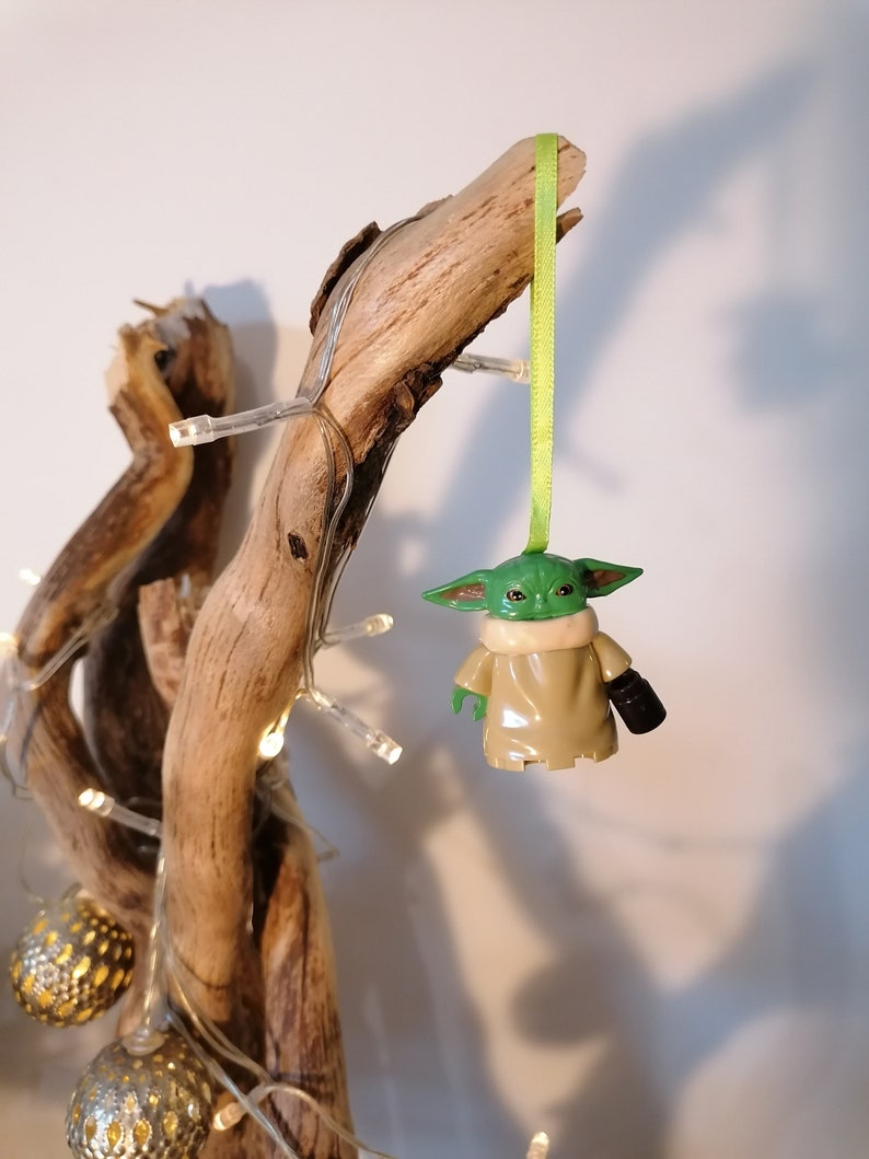 May include: A green and brown Yoda ornament hanging from a branch with a green ribbon. The ornament is made of plastic and has a detailed design.