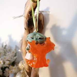 May include: A green and orange Christmas bauble featuring a character from the Star Wars franchise. The character is holding a small orange tree.
