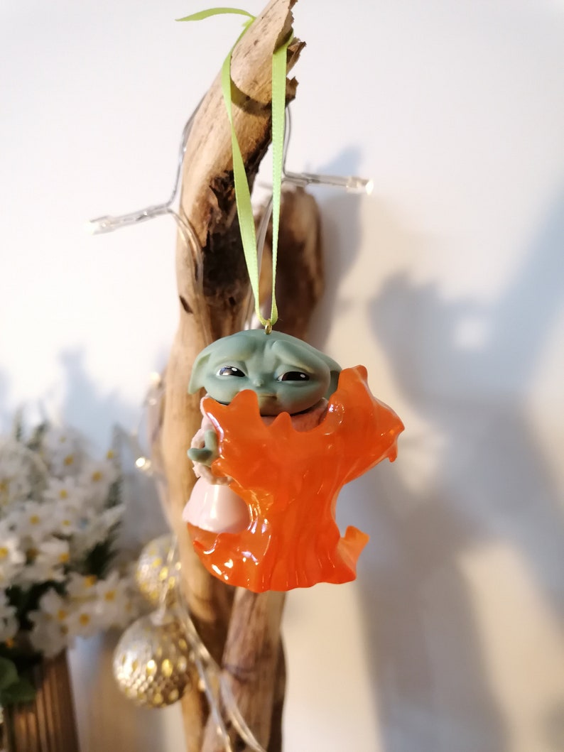 May include: A green and orange Christmas bauble featuring a character from the Star Wars franchise. The character is holding a small orange tree.