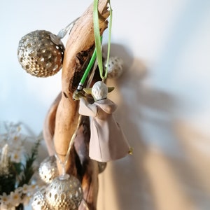 Puede incluir: Adorno navide&ntilde;o de Yoda con un sable de luz verde y una t&uacute;nica beige colgado de un trozo de madera flotante. El adorno est&aacute; suspendido por una cinta verde. Luces de globo plateadas y flores blancas tambi&eacute;n son visibles.