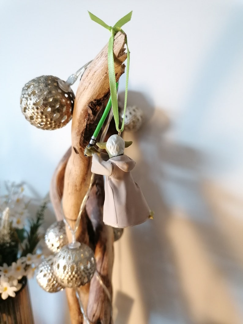 Puede incluir: Adorno navide&ntilde;o de Yoda con un sable de luz verde y una t&uacute;nica beige colgado de un trozo de madera flotante. El adorno est&aacute; suspendido por una cinta verde. Luces de globo plateadas y flores blancas tambi&eacute;n son visibles.