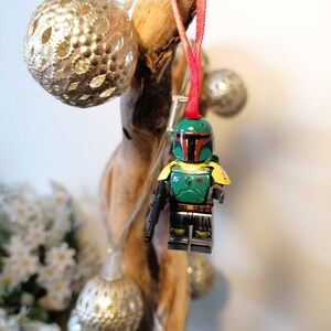 May include: A green and yellow Lego minifigure of Boba Fett hanging from a branch with a red ribbon. The minifigure is holding a blaster rifle and has a green helmet with a red visor.