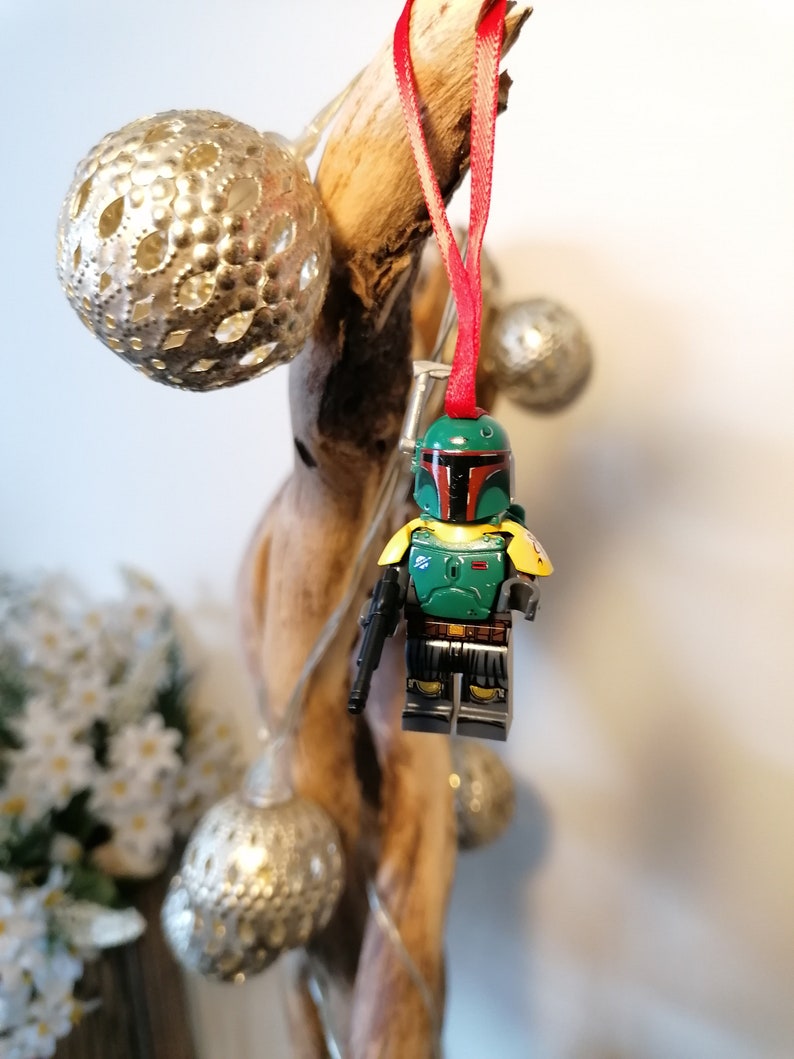May include: A green and yellow Lego minifigure of Boba Fett hanging from a branch with a red ribbon. The minifigure is holding a blaster rifle and has a green helmet with a red visor.