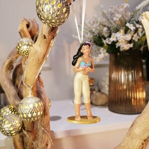 May include: A white string hangs from a branch with a small figurine of a woman with brown hair and a blue top. The figurine is holding a flower. The branch is decorated with small, round, silver ornaments with holes in them.