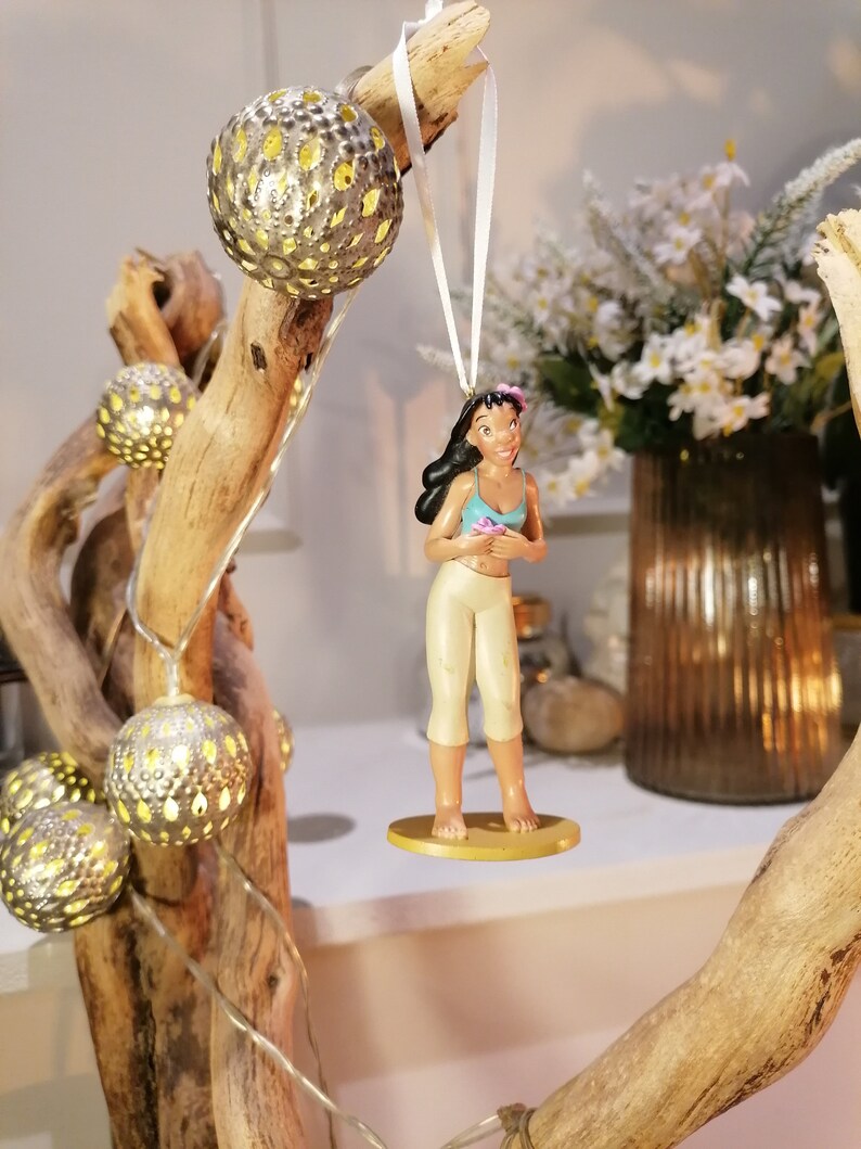 May include: A white string hangs from a branch with a small figurine of a woman with brown hair and a blue top. The figurine is holding a flower. The branch is decorated with small, round, silver ornaments with holes in them.