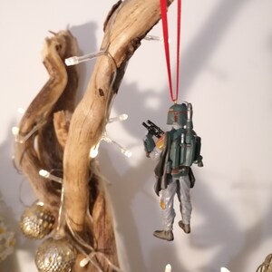 May include: A green and brown Star Wars Boba Fett ornament hanging from a branch with a red ribbon. The ornament is hanging from a branch with white lights wrapped around it.