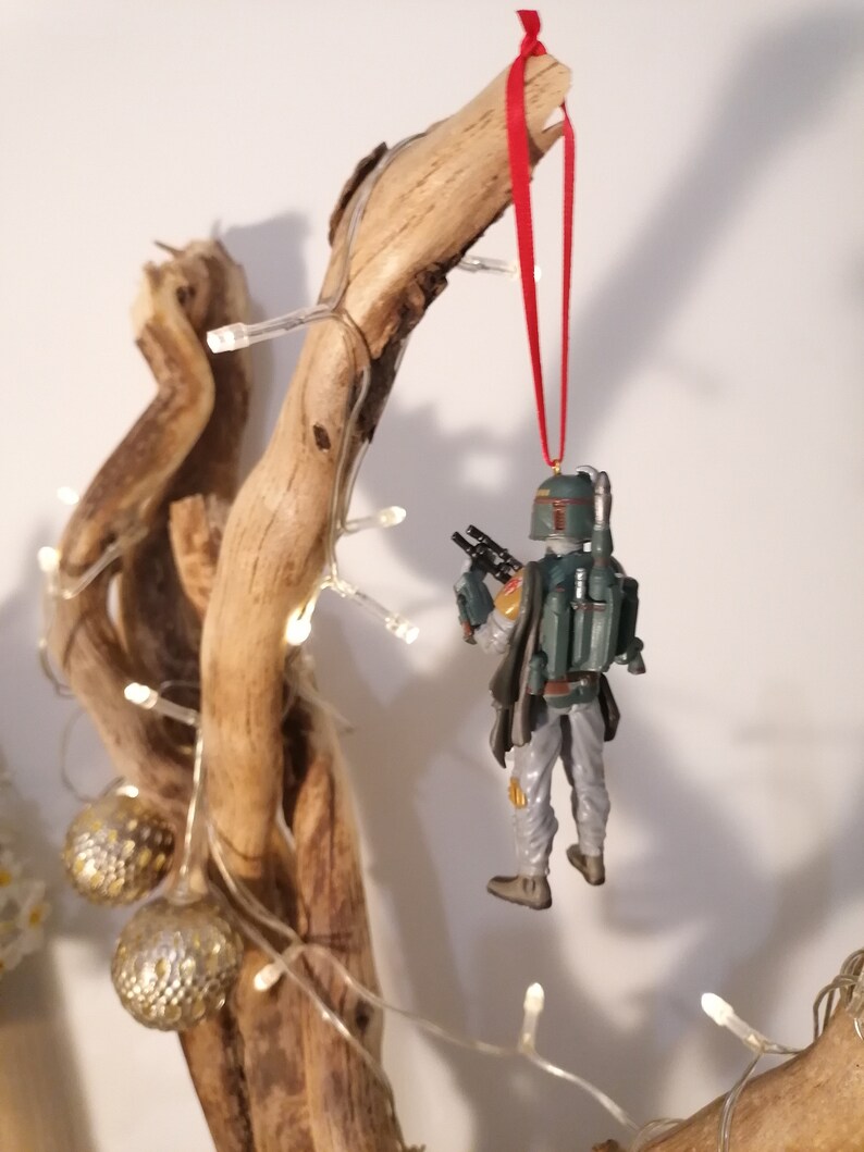 May include: A green and brown Star Wars Boba Fett ornament hanging from a branch with a red ribbon. The ornament is hanging from a branch with white lights wrapped around it.