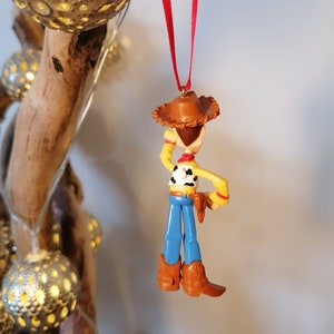 Pu&ograve; includere: Uno spago rosso pende dalla cima di un ramo di legno con una statuetta di plastica di Woody di Toy Story appesa allo spago. Woody indossa il suo cappello marrone distintivo, jeans blu e un gilet giallo con una bandana rossa. Tiene la sua pistola nella mano destra.