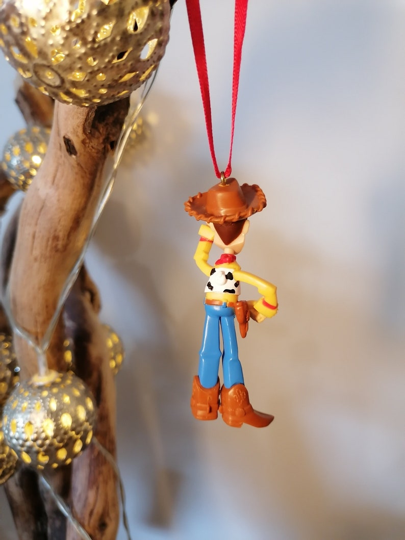Pu&ograve; includere: Uno spago rosso pende dalla cima di un ramo di legno con una statuetta di plastica di Woody di Toy Story appesa allo spago. Woody indossa il suo cappello marrone distintivo, jeans blu e un gilet giallo con una bandana rossa. Tiene la sua pistola nella mano destra.