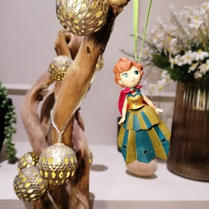Pu&ograve; includere: Una piccola statuina di plastica di una principessa dai capelli rossi che indossa un abito dorato e turchese e un mantello rosso. La statuina &egrave; appesa a un nastro verde su un ramo con lampadine decorative dorate.