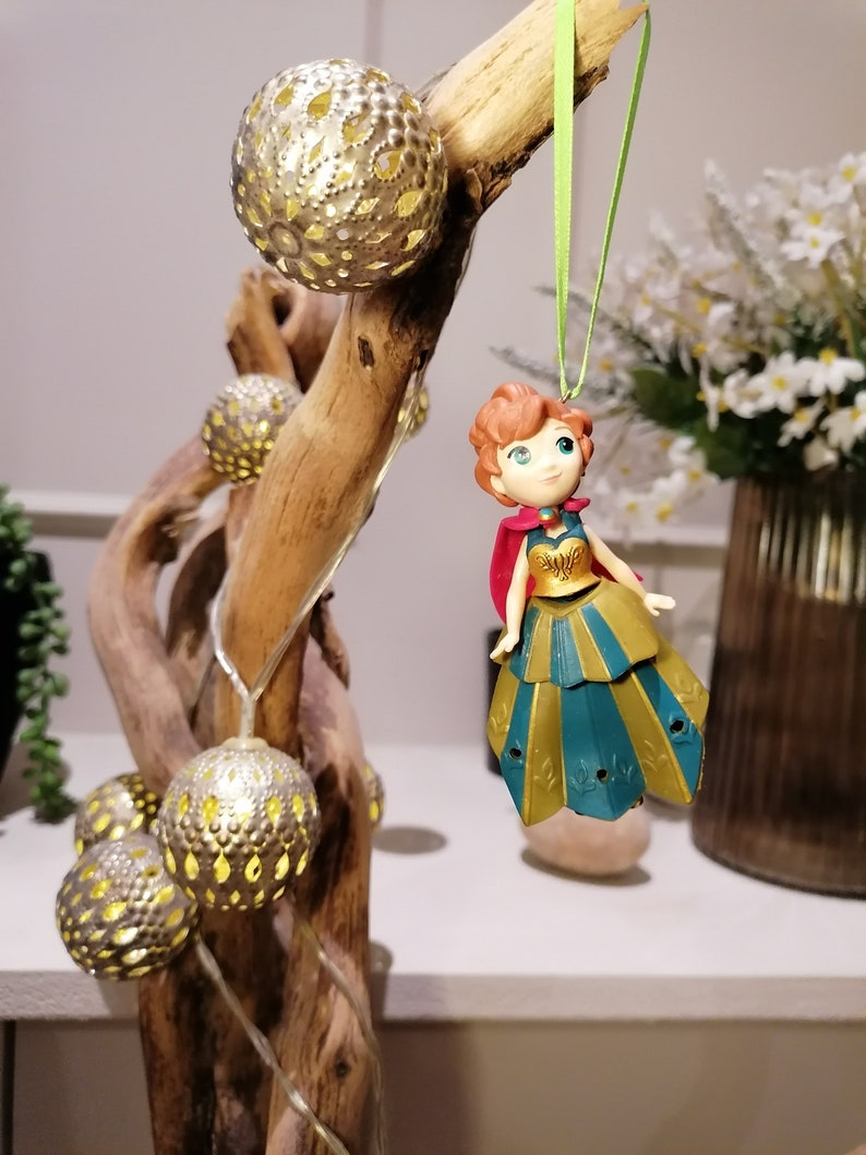 Pu&ograve; includere: Una piccola statuina di plastica di una principessa dai capelli rossi che indossa un abito dorato e turchese e un mantello rosso. La statuina &egrave; appesa a un nastro verde su un ramo con lampadine decorative dorate.