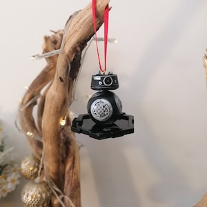 K&ouml;nnte beinhalten: Schwarzer und silberner Star Wars BB-8 Weihnachtsbaumschmuck mit einem roten Band, das an einem Ast h&auml;ngt.