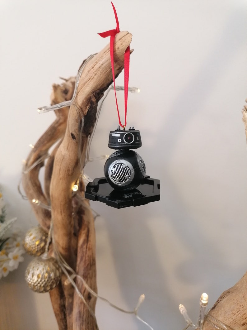 K&ouml;nnte beinhalten: Schwarzer und silberner Star Wars BB-8 Weihnachtsbaumschmuck mit einem roten Band, das an einem Ast h&auml;ngt.