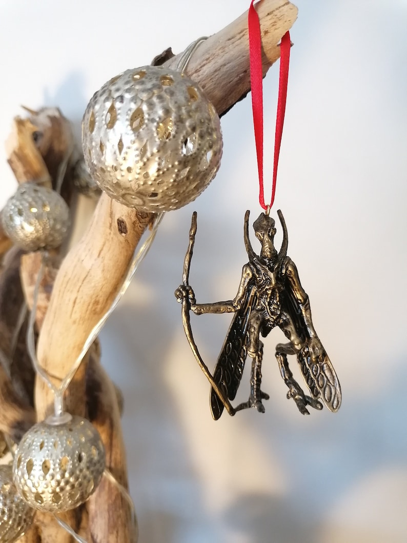 May include: A bronze-coloured ornament of a winged creature with a bow and arrow. The ornament is hanging from a red ribbon on a branch with silver and white ornaments.