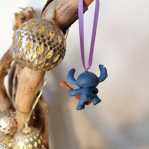 Op de afbeelding: Blauw Stitch-personage ornament dat gitaar speelt, hangend aan een paars lint aan een tak met gouden kerstverlichting.
