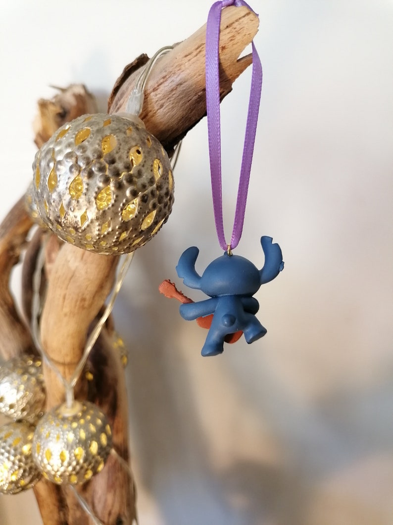 Op de afbeelding: Blauw Stitch-personage ornament dat gitaar speelt, hangend aan een paars lint aan een tak met gouden kerstverlichting.