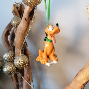 Pode incluir: Adorno de Natal de plástico laranja e castanho do Pluto, o cão da Disney, segurando um osso branco. O adorno está pendurado numa fita verde.