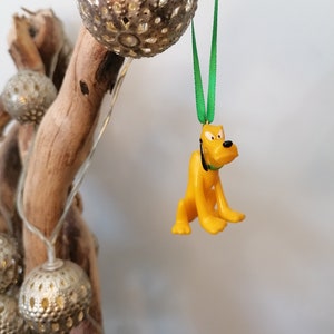 Op de afbeelding: Een gele plastic ornament van Pluto, een cartoonhond van Disney, hangend aan een groen lint. De ornament hangt aan een tak met zilveren decoratieve ballen.