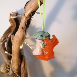 May include: A green and orange Christmas bauble featuring a character from the Star Wars franchise. The character is holding a small orange object in its hands.