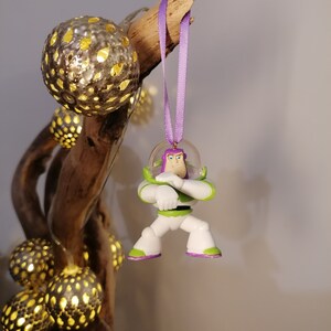 Puede incluir: Un adorno de plástico morado y verde del personaje Buzz Lightyear de la franquicia Toy Story. El adorno está colgado de una cinta morada.