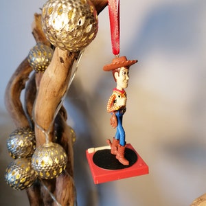 Puede incluir: Un adorno navide&ntilde;o rojo y blanco con una figura de Woody de la pel&iacute;cula Toy Story. Woody lleva su caracter&iacute;stico sombrero de vaquero, chaleco y pantalones vaqueros azules. Est&aacute; de pie sobre una plataforma roja con un tocadiscos negro.