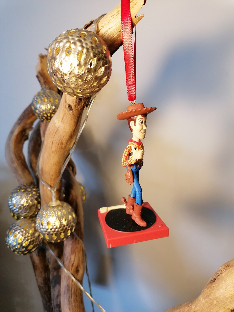 Puede incluir: Un adorno navide&ntilde;o rojo y blanco con una figura de Woody de la pel&iacute;cula Toy Story. Woody lleva su caracter&iacute;stico sombrero de vaquero, chaleco y pantalones vaqueros azules. Est&aacute; de pie sobre una plataforma roja con un tocadiscos negro.
