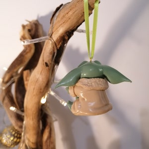 May include: A green and brown figurine of a character from the Star Wars franchise, hanging from a green ribbon on a branch of driftwood. The figurine is wearing a brown coat and has large ears.